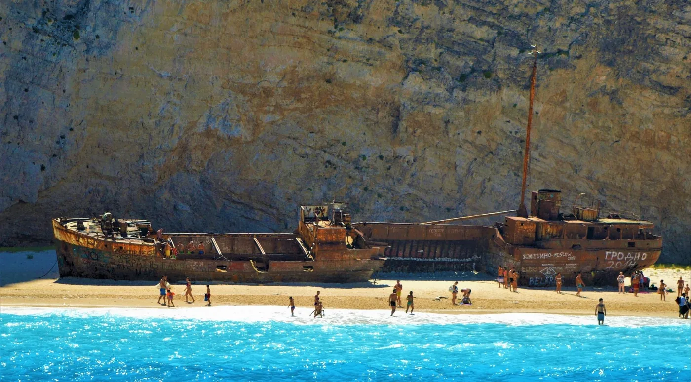 Visitors walking on the sand beside the rusted hull of the Panagiotis shipwreck at Navagio Beach in an earlier season