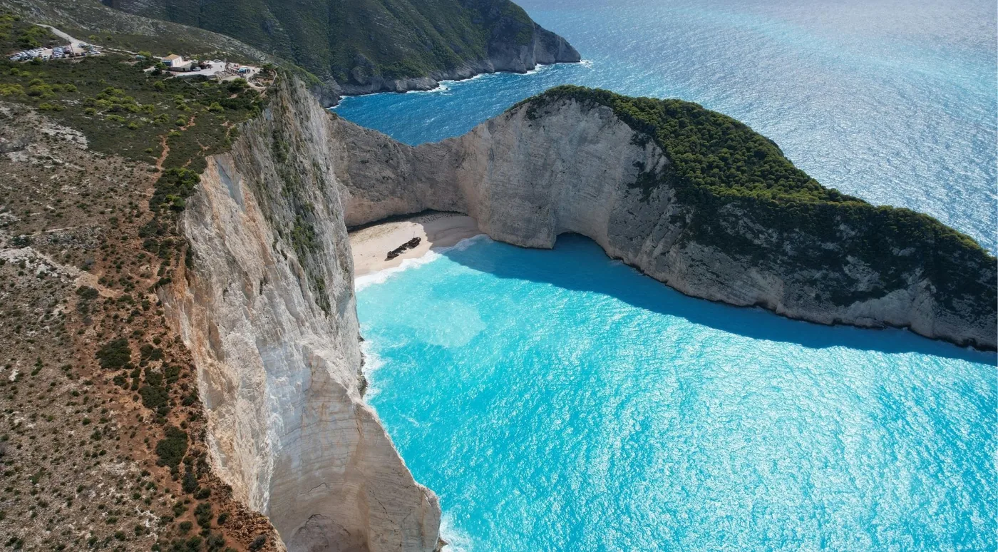 High aerial view of Navagio cove with turquoise shallows meeting deep blue water below sheer limestone cliffs