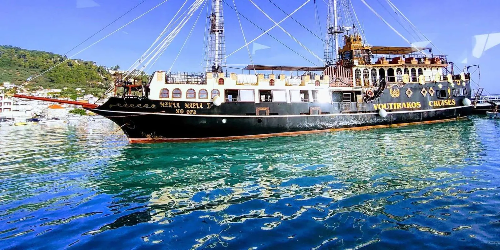 A dark-hulled pirate ship with rigging and masts moored on calm water below a wooded shore