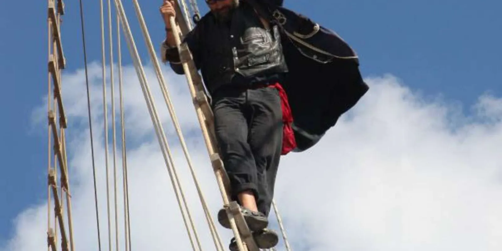 A costumed crew member climbing the rigging against a bright cloudy sky