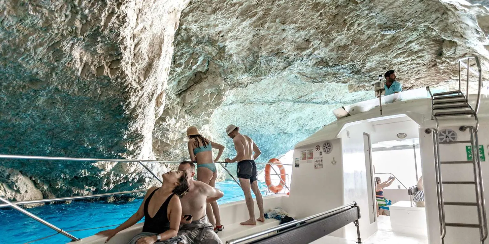 Passengers on the open deck of a small boat inside a sea cave, with sunlit turquoise water alongside