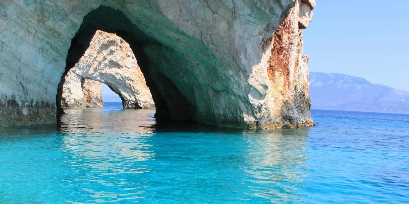 A natural stone arch rising from clear turquoise water, with mountains on the horizon beyond