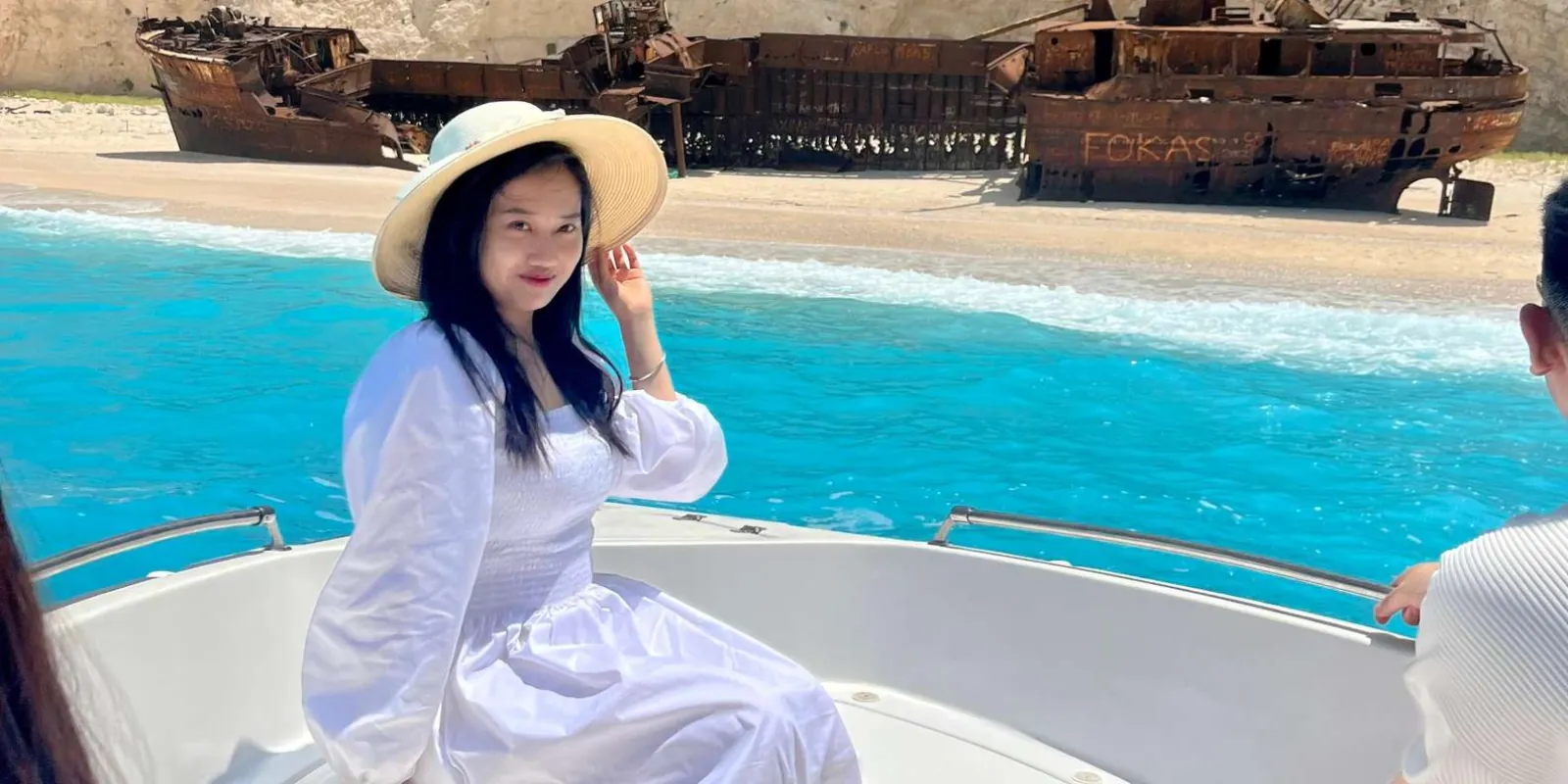 A visitor in a white dress on the deck of a boat, with the rusted shipwreck on the sand behind
