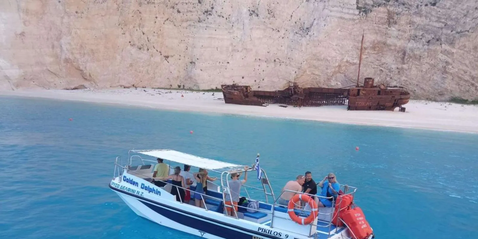 A small tour boat with passengers holding off the beach, with the rusted shipwreck on the sand behind